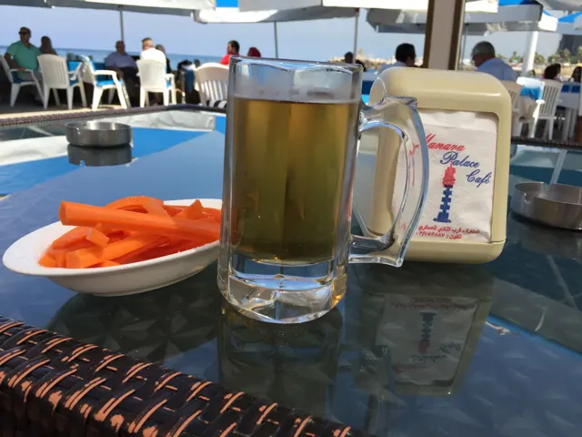 A glass mug of a golden drink stands beside a bowl of carrot sticks on a reflective outdoor table.