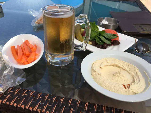 A glass of a golden drink, carrot sticks, vegetables and a pale dip are arranged on an outdoor table.