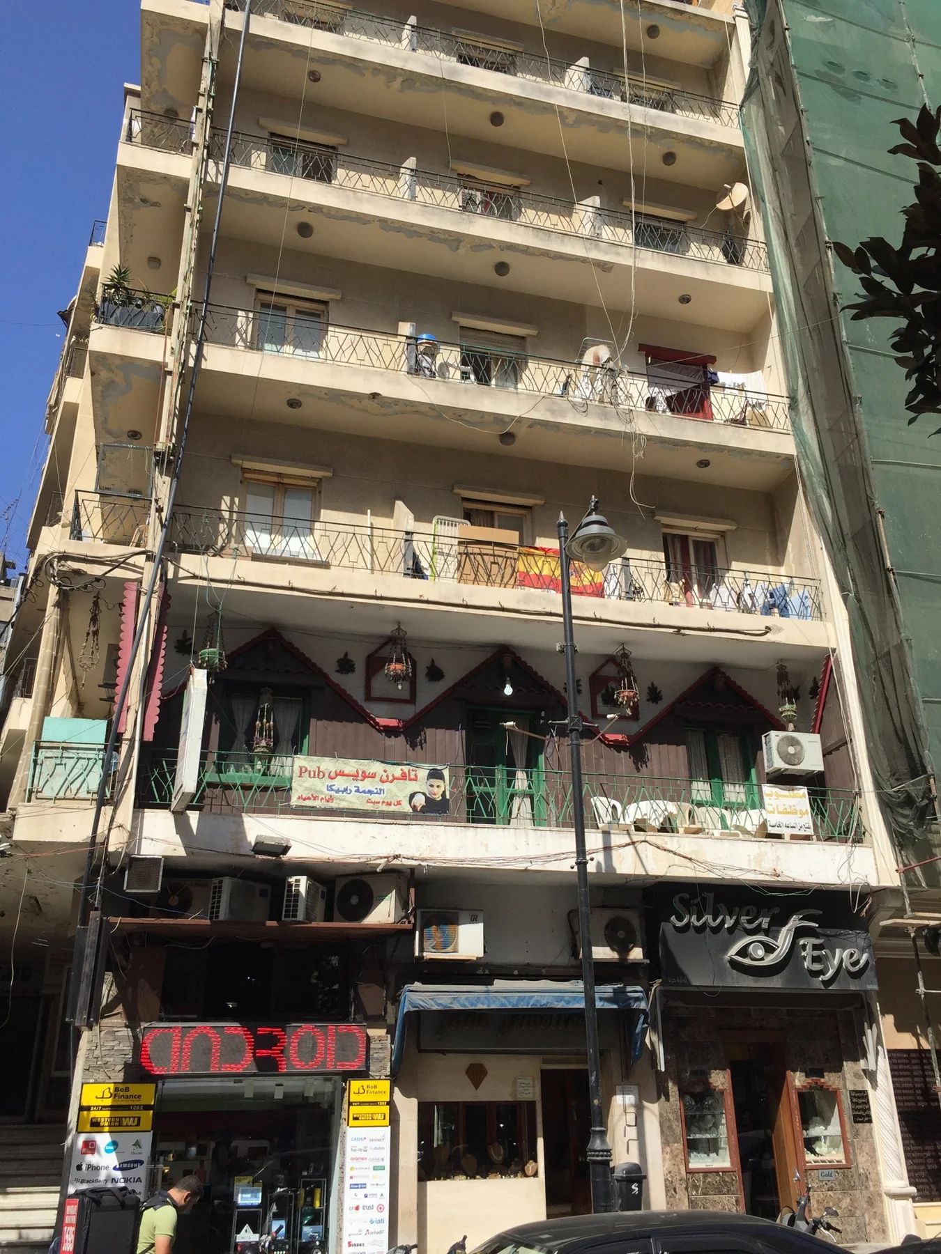 A portrait view follows a weathered apartment facade with projecting balconies above street-level shops.