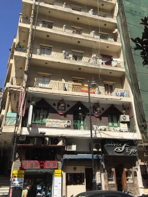 A portrait view follows a weathered apartment facade with projecting balconies above street-level shops.