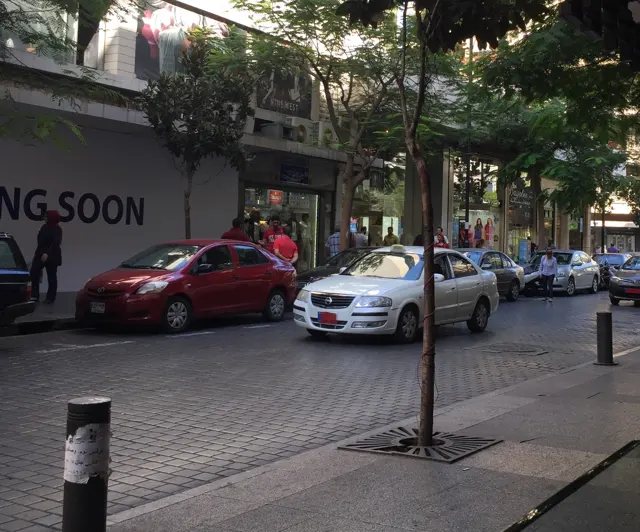 Red and white cars stand along a tree-lined street beside shopfronts and short bollards.
