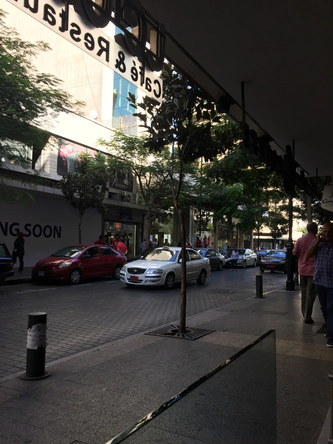 An awning frames a shaded pavement, parked cars and trees outside street-level shops.