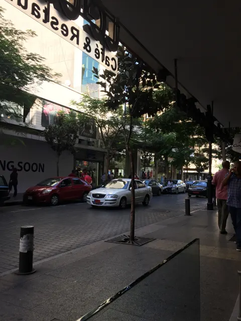 An awning frames a shaded pavement, parked cars and trees outside street-level shops.