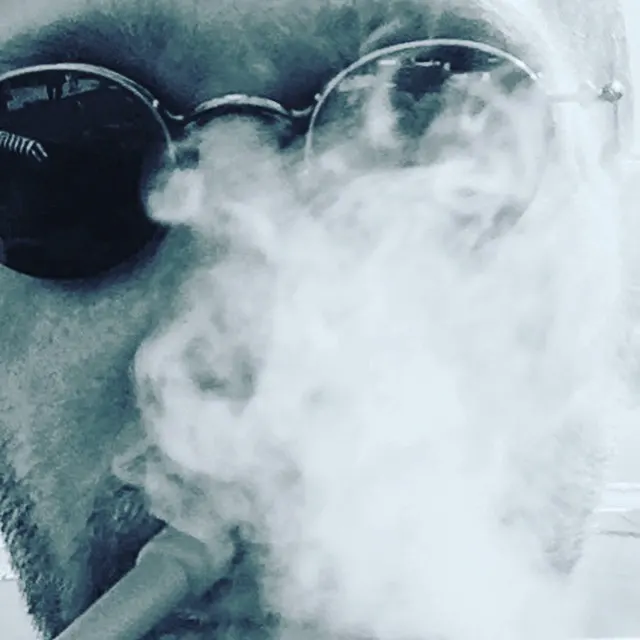 A close-up of a face wearing dark sunglasses is partly obscured by a pale cloud of smoke.