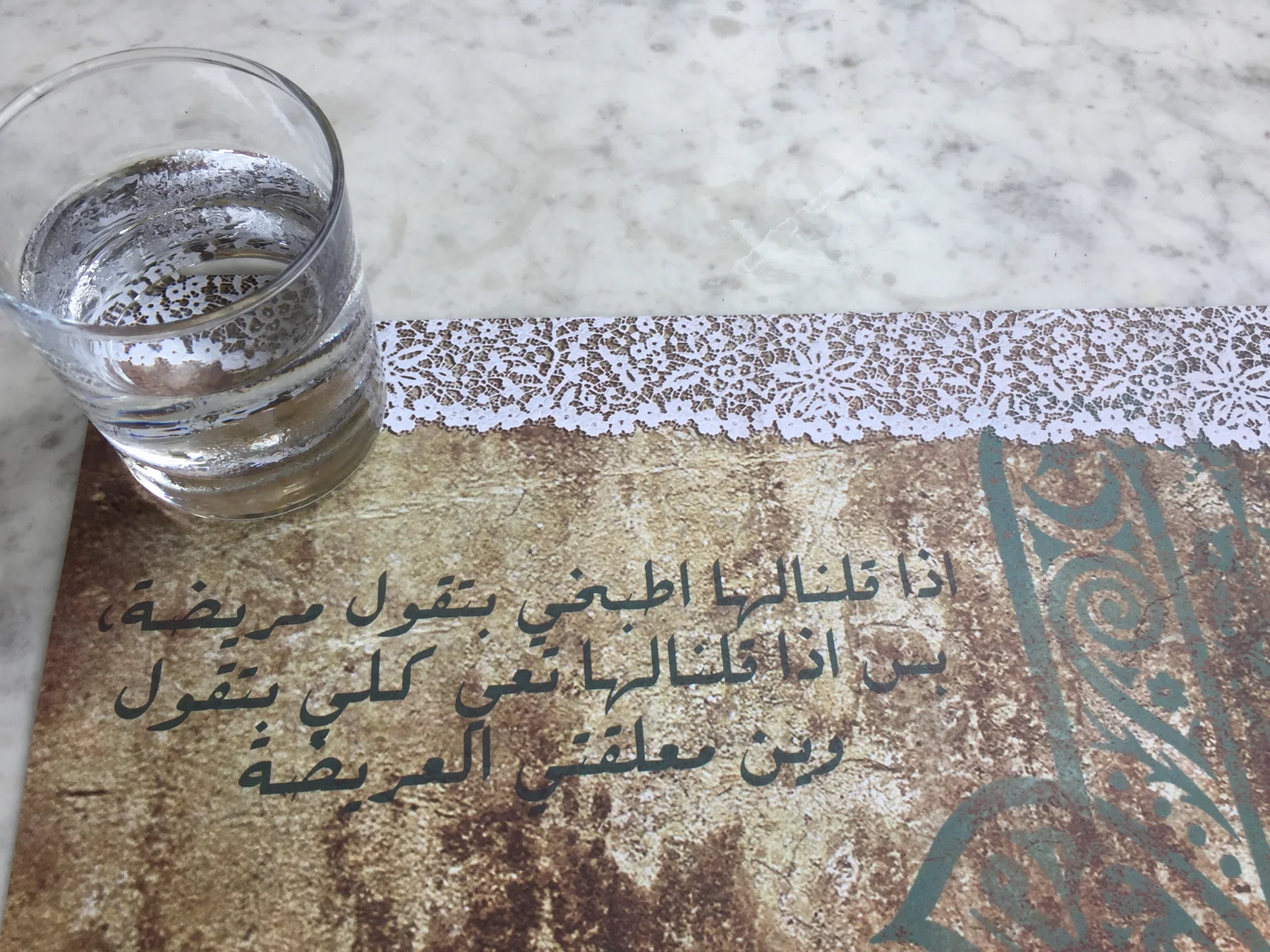 A water glass rests on a tabletop beside decorative Arabic lettering and a patterned border.