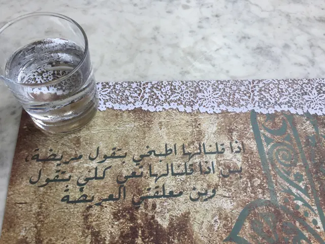 A water glass rests on a tabletop beside decorative Arabic lettering and a patterned border.