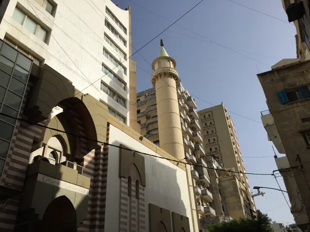 An upward street view shows a minaret squeezed between tall buildings and overhead cables.