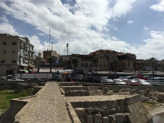 A stone walkway and low masonry lead towards parked cars and waterfront buildings beneath a cloudy sky.