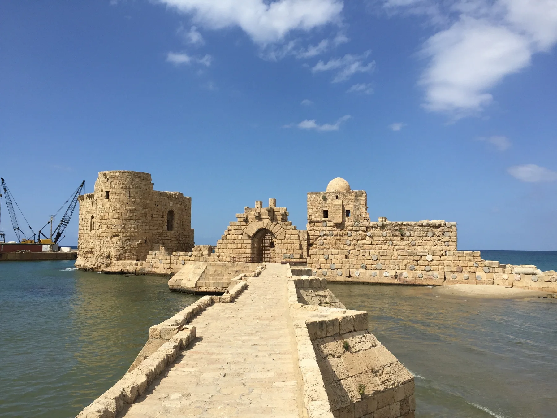 A narrow stone causeway crosses blue-green water towards the towers and arched entrance of Sidon Sea Castle.