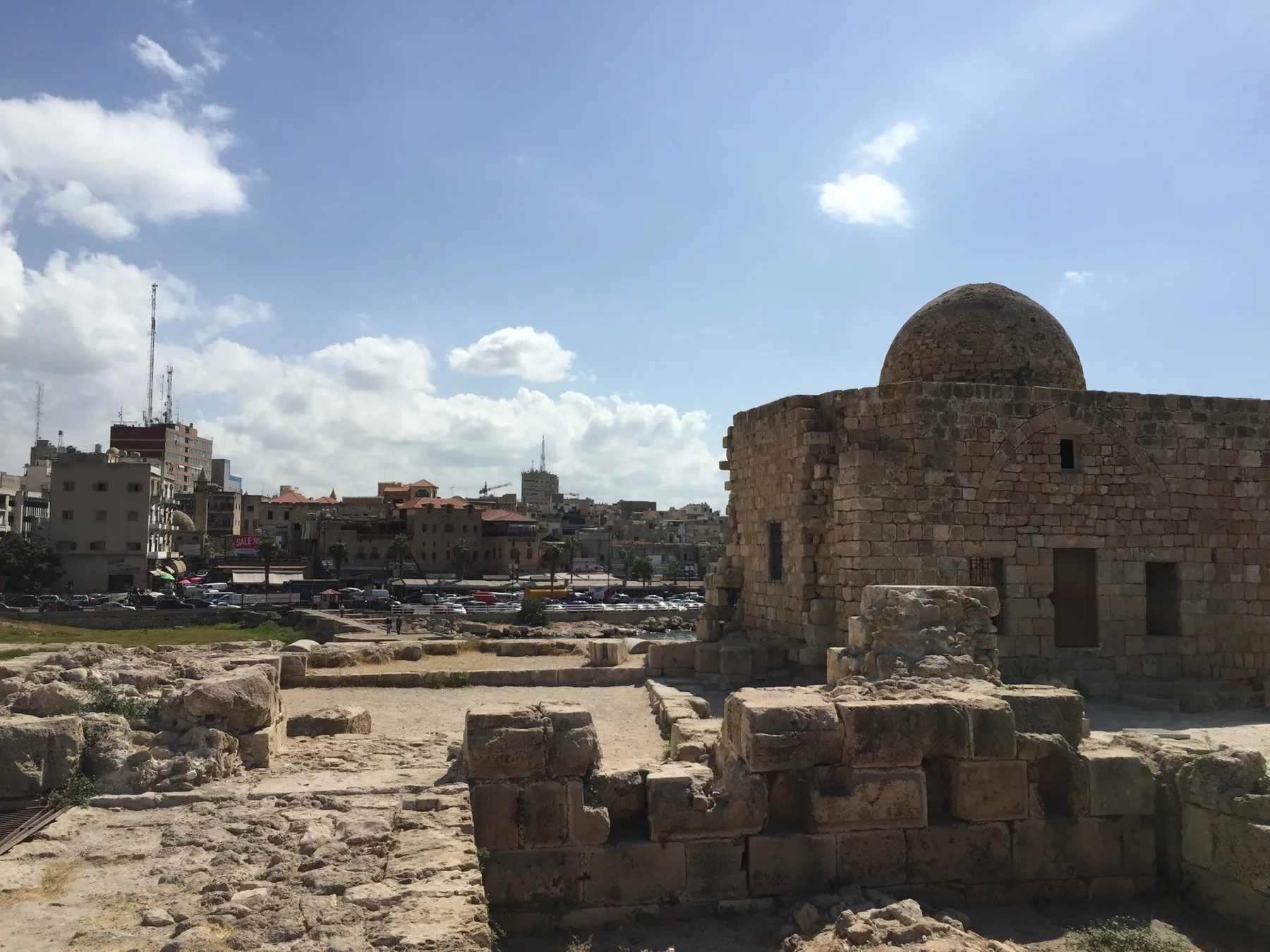 A small domed stone structure stands beside low ruined walls, with the waterfront town behind.