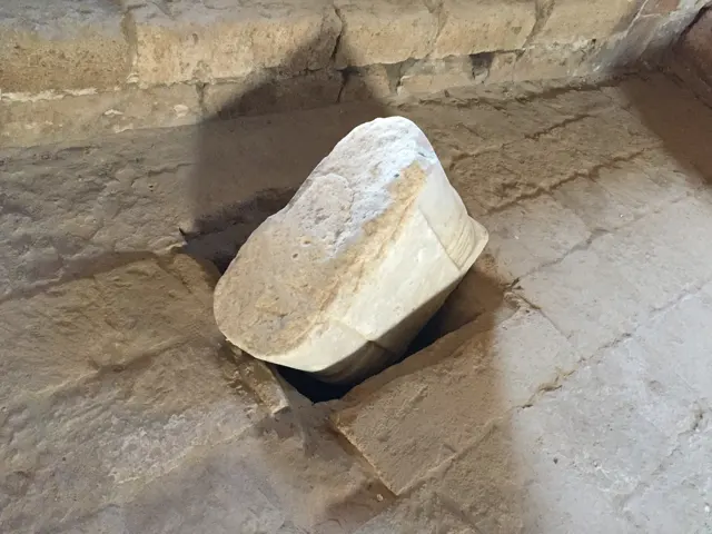 A thick circular stone piece rests at an angle within an opening in a worn stone floor.