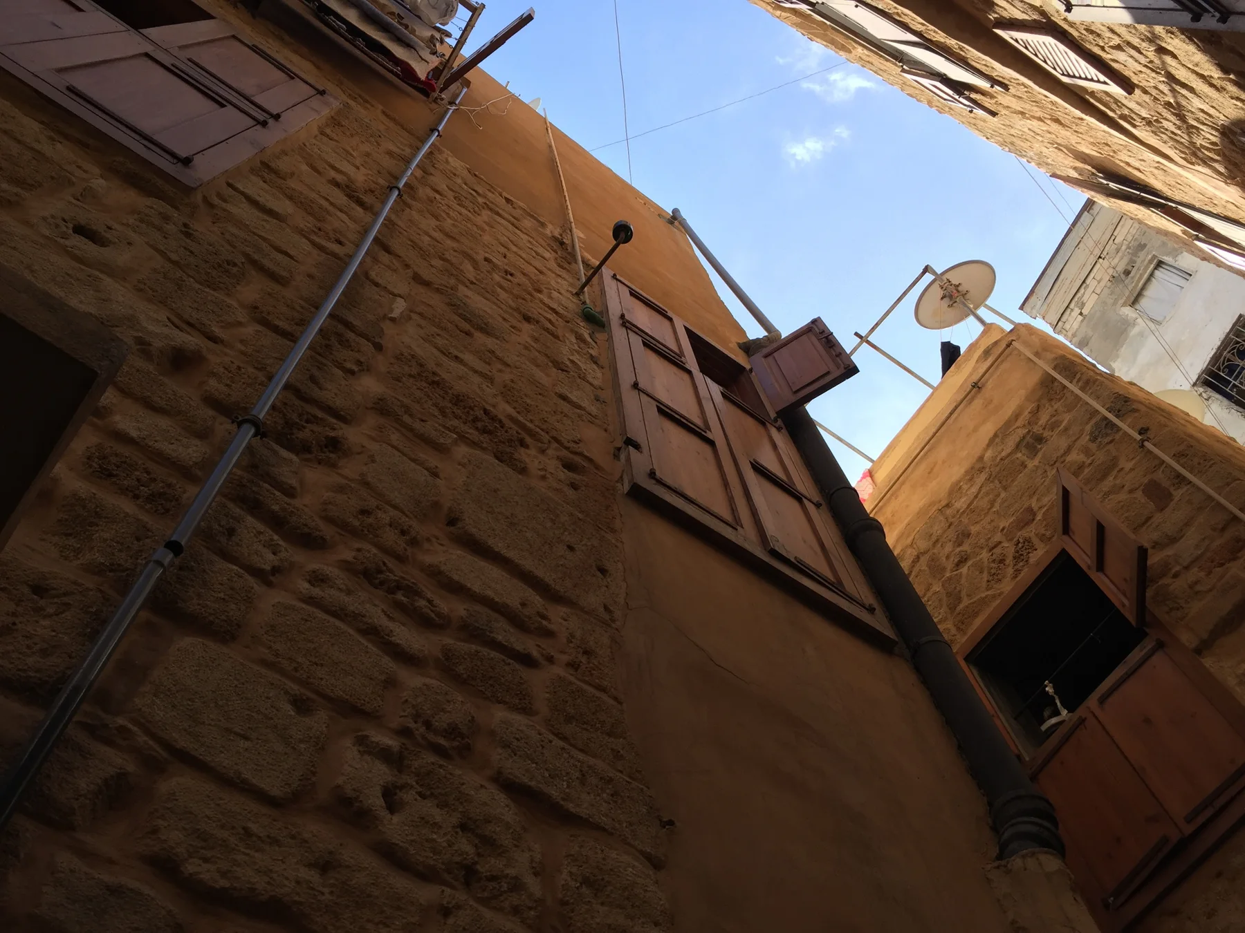 An upward view between closely spaced stone buildings shows wooden shutters, cables and a patch of blue sky.