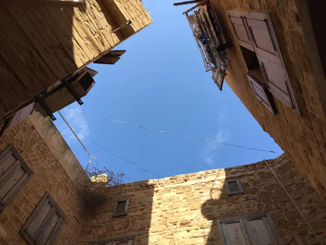 Stone walls with wooden shutters frame a small opening of blue sky above a narrow passage.