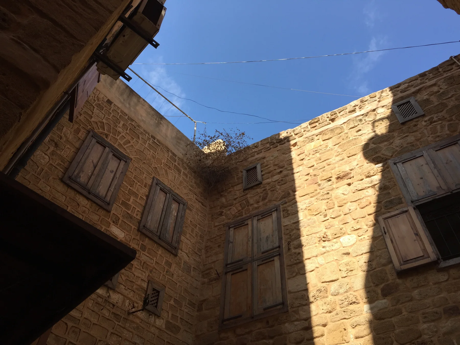 Warm stone walls and shuttered windows rise around a narrow courtyard beneath bright blue sky.