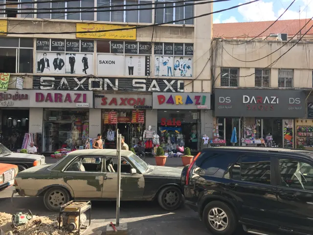 Cars pass rows of shopfronts and large signs beneath an apartment building.