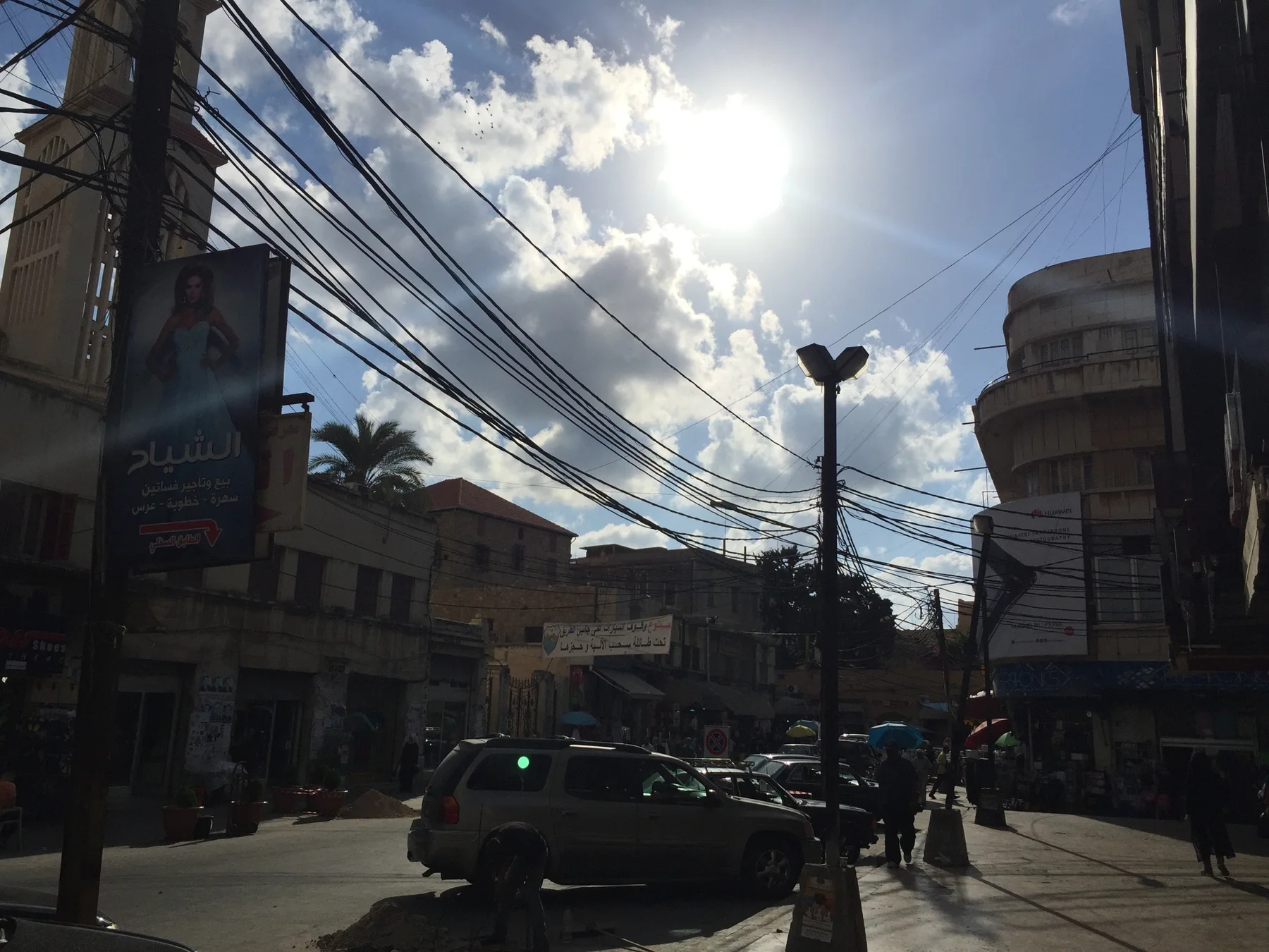 Dense overhead wires cross a sunlit street between buildings, with cars and pedestrians below.