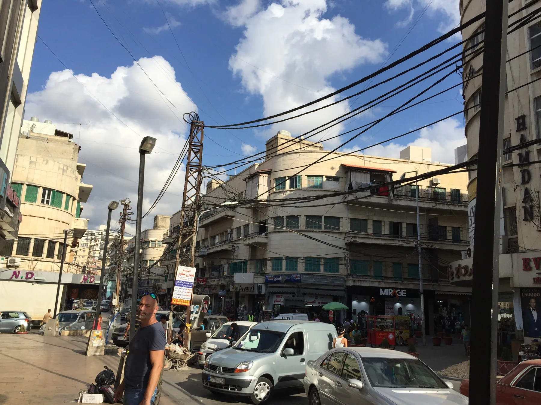 Vehicles gather at a busy junction beneath tangled utility wires and a bright clouded sky.