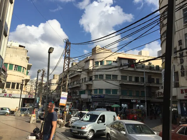 Vehicles gather at a busy junction beneath tangled utility wires and a bright clouded sky.