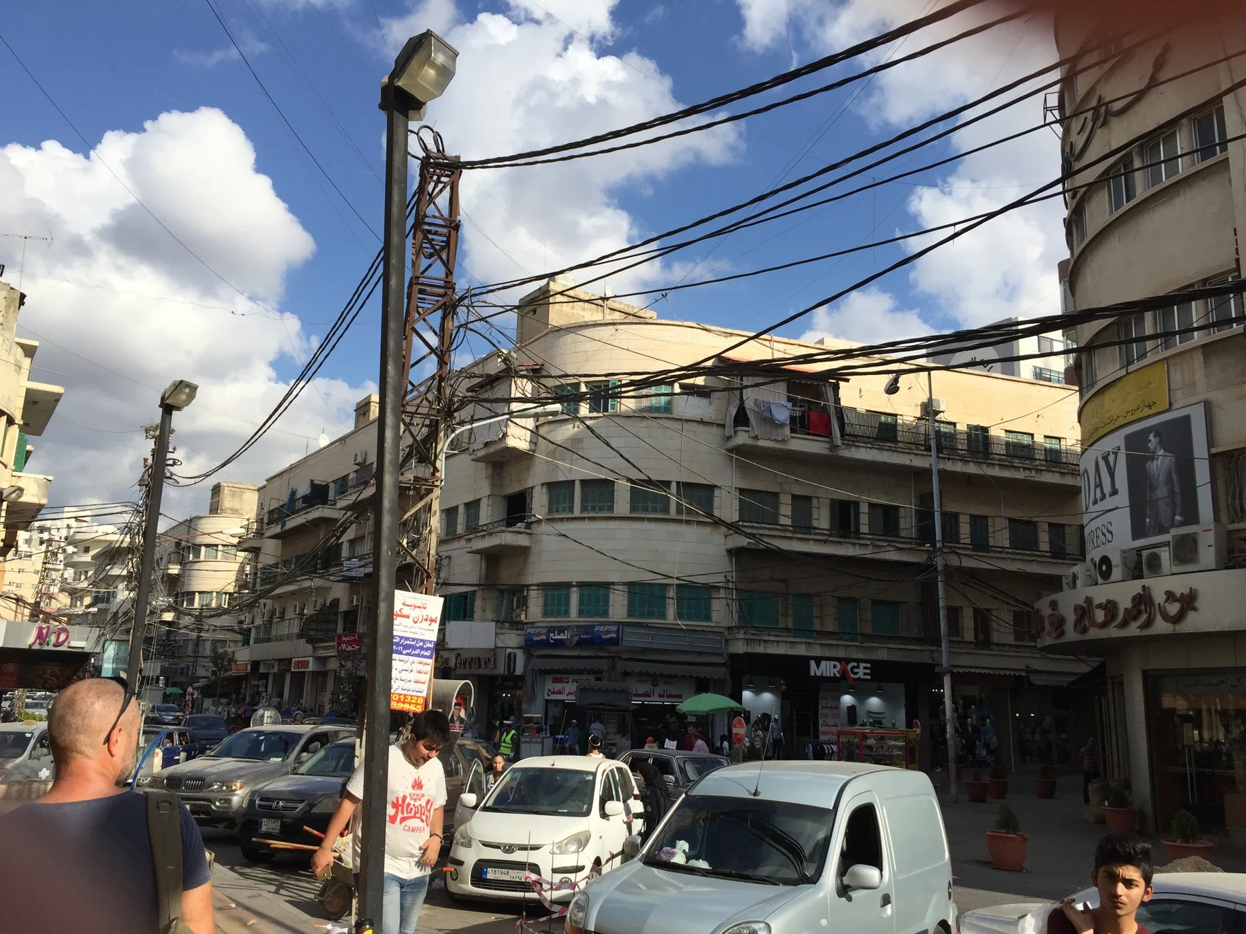 A tall pole carrying numerous cables rises above traffic, shops and pedestrians.