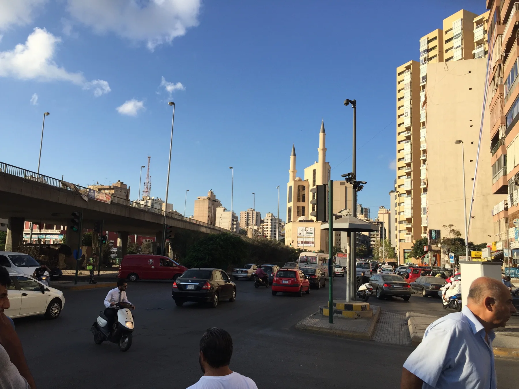 Cars and pedestrians share a broad city road between tall buildings, with slender minarets in the distance.