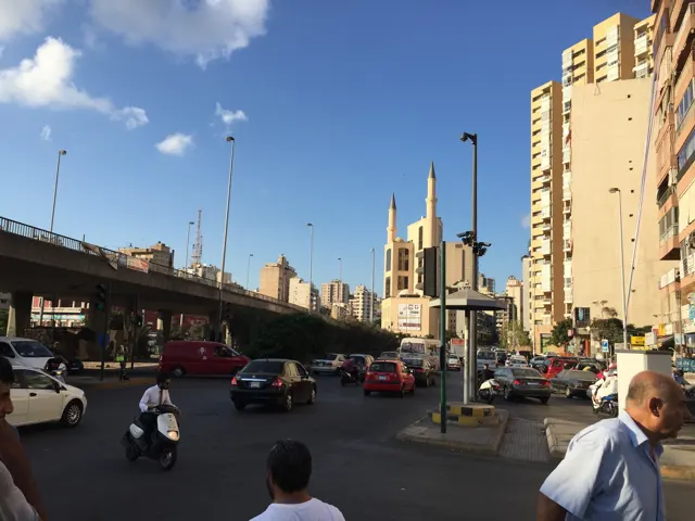Cars and pedestrians share a broad city road between tall buildings, with slender minarets in the distance.