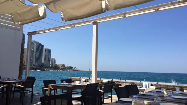 White canopy supports frame outdoor tables and blue sea, with high-rises along the shore to the left.