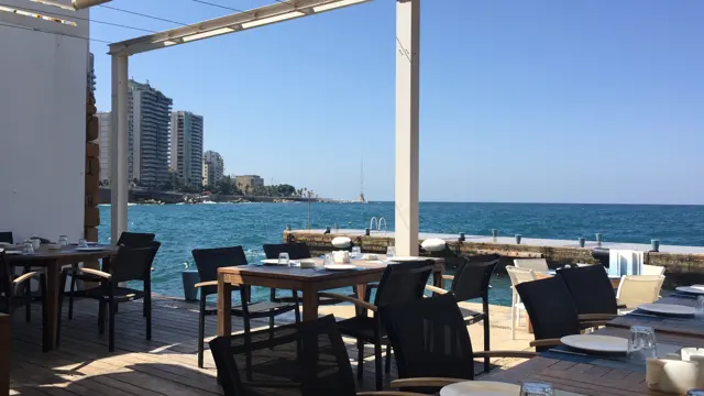 Tables and dark chairs face blue water beneath a terrace canopy, with distant seafront buildings.