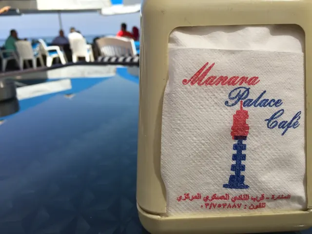 A napkin holder printed with a cafe name and lighthouse motif stands on an outdoor table.
