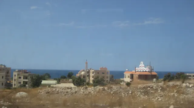 Scattered buildings and open ground lie between the roadside and a distant blue sea.