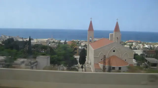 A pale church with two red-roofed spires stands among low buildings with the sea beyond.