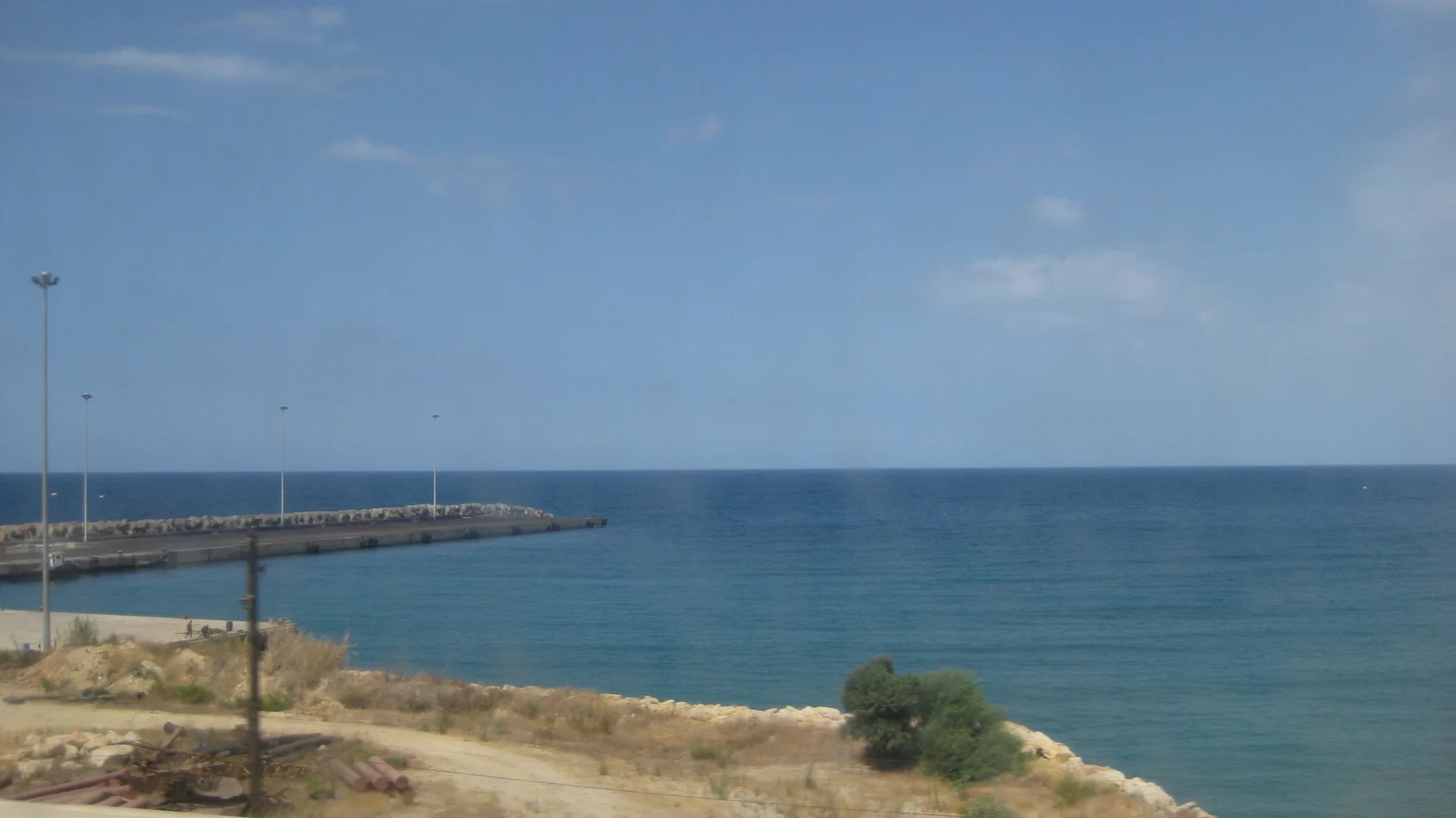 A long breakwater projects into deep blue water beside a rocky stretch of coast.