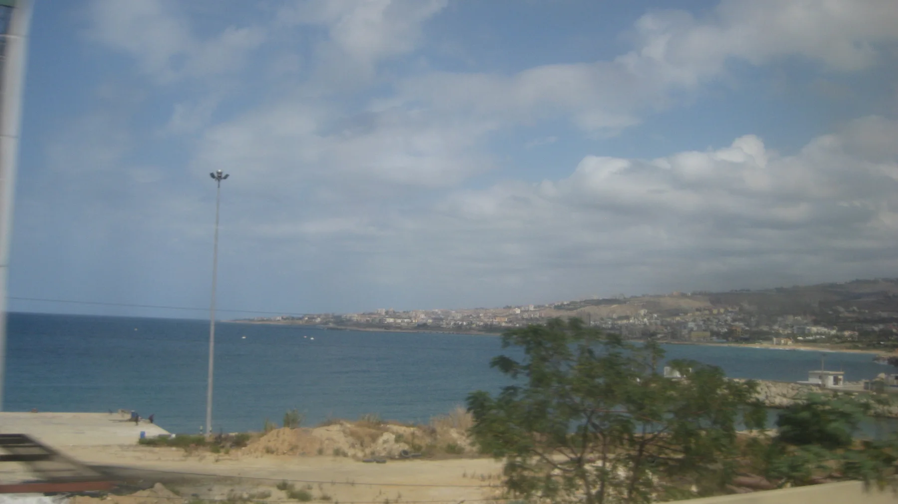 A rocky foreground overlooks blue water and a curving developed shoreline beneath scattered clouds.