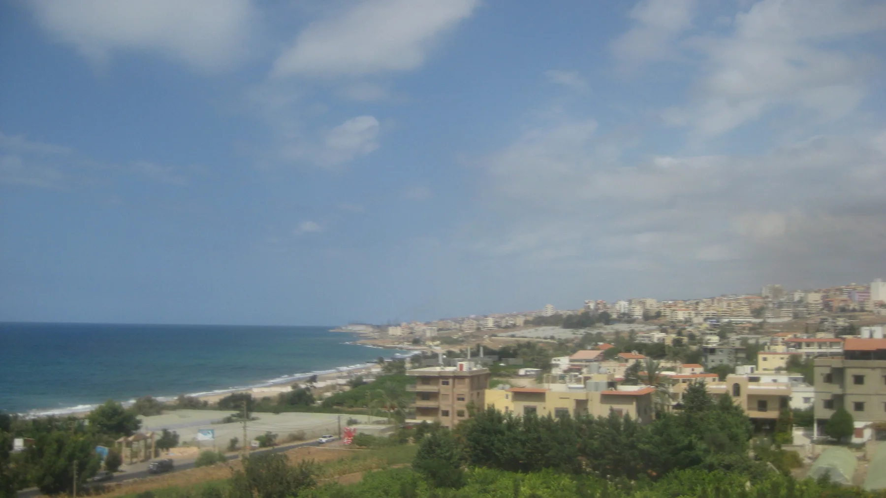 A dense coastal town follows a curving beach and blue bay in an elevated view.