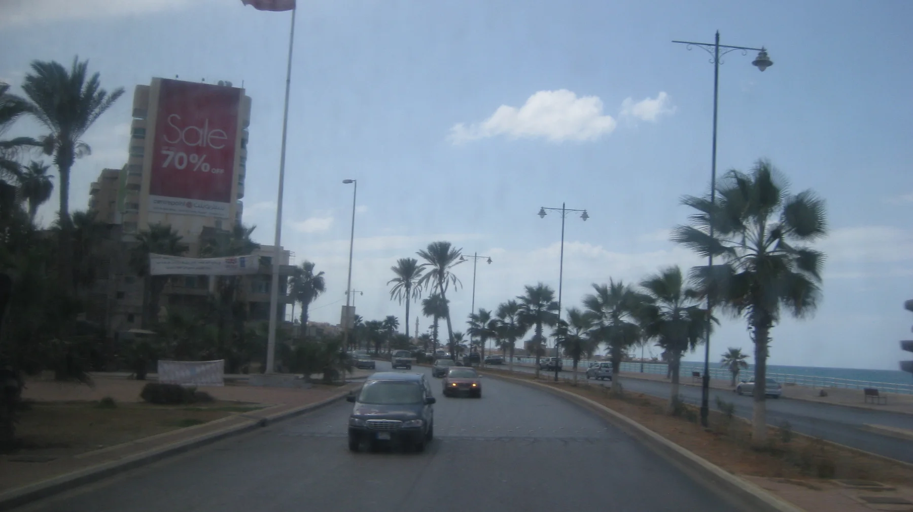 Cars follow a coastal road lined with palms and street lamps beside the sea.