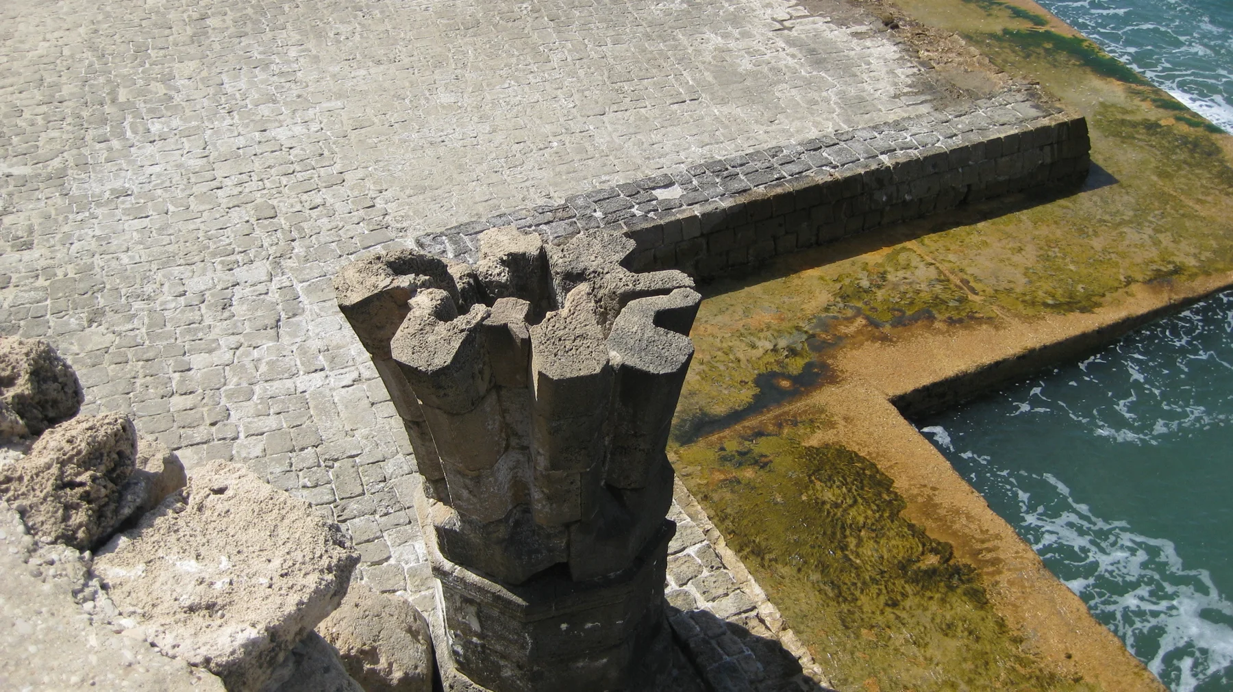 A weathered stone post rises above a paved platform edged with shallow water and seaweed.