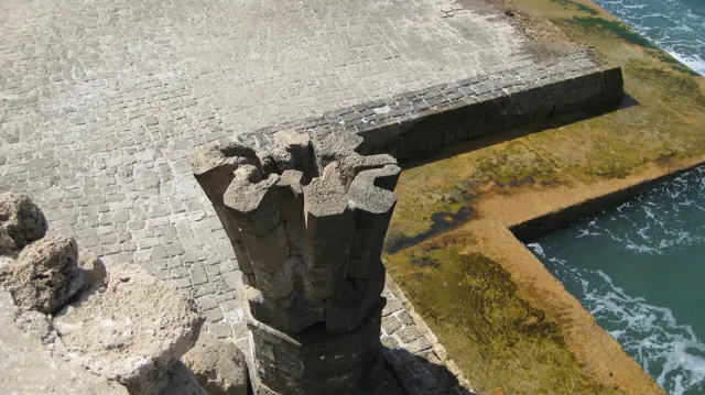 A weathered stone post rises above a paved platform edged with shallow water and seaweed.