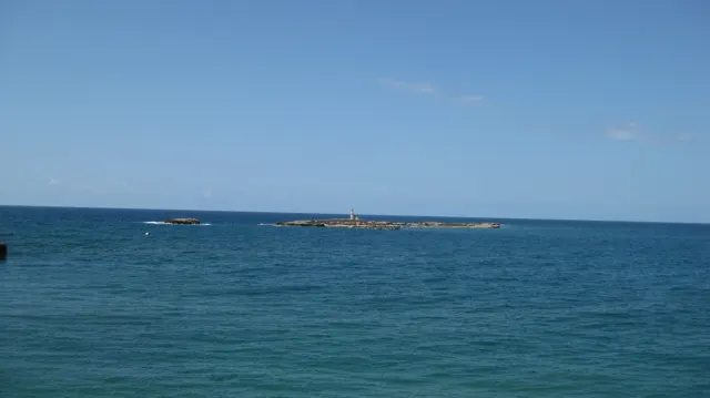 Open blue sea stretches towards low rocks near the horizon beneath a clear sky.
