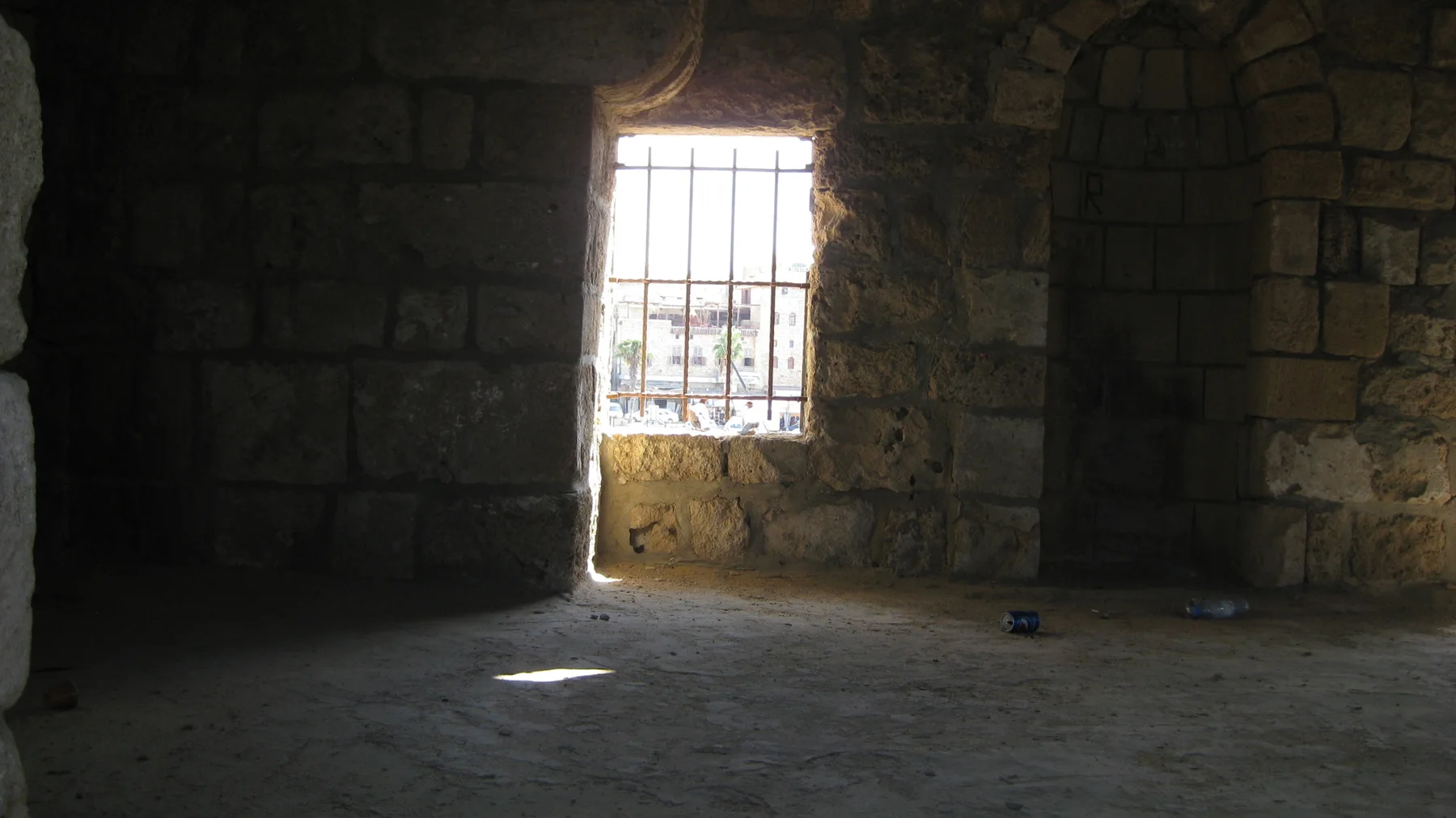 A barred window admits a bright rectangle of light into a dark stone room.