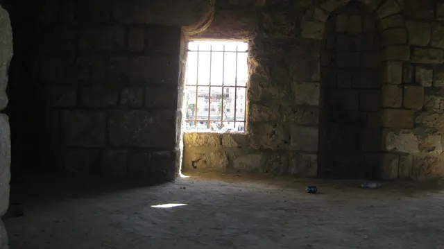 A barred window admits a bright rectangle of light into a dark stone room.
