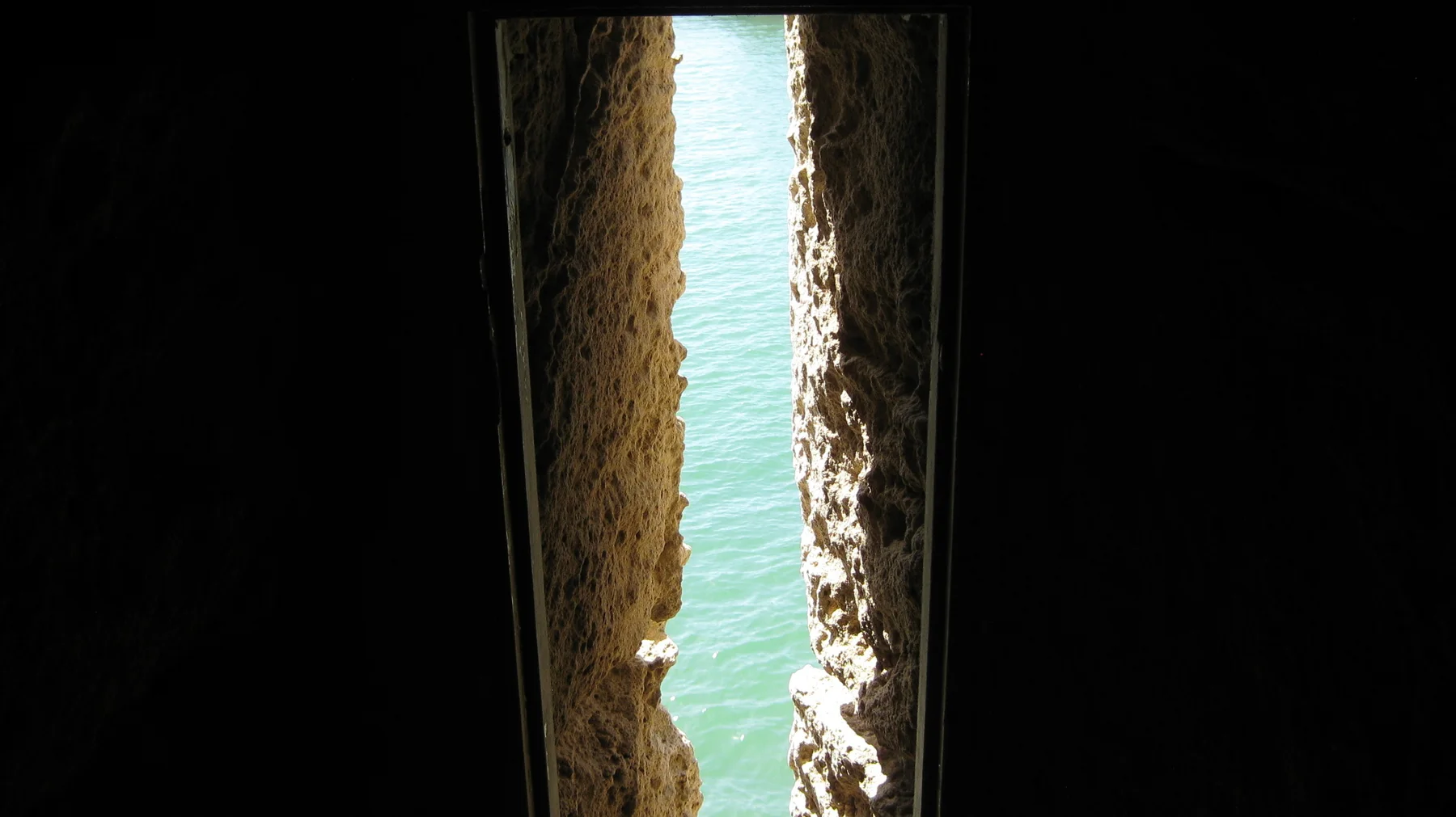 A thin opening between rough dark stone walls reveals bright turquoise water outside.