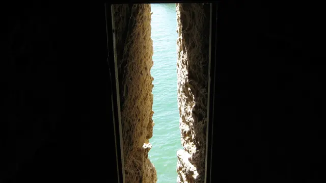 A thin opening between rough dark stone walls reveals bright turquoise water outside.