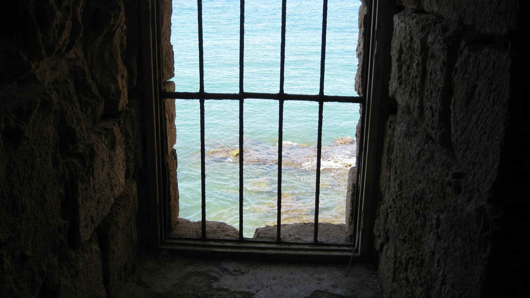 Iron bars divide a narrow window looking out from a dark stone wall towards blue-green water.