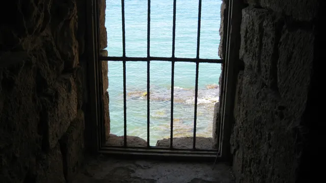 Iron bars divide a narrow window looking out from a dark stone wall towards blue-green water.