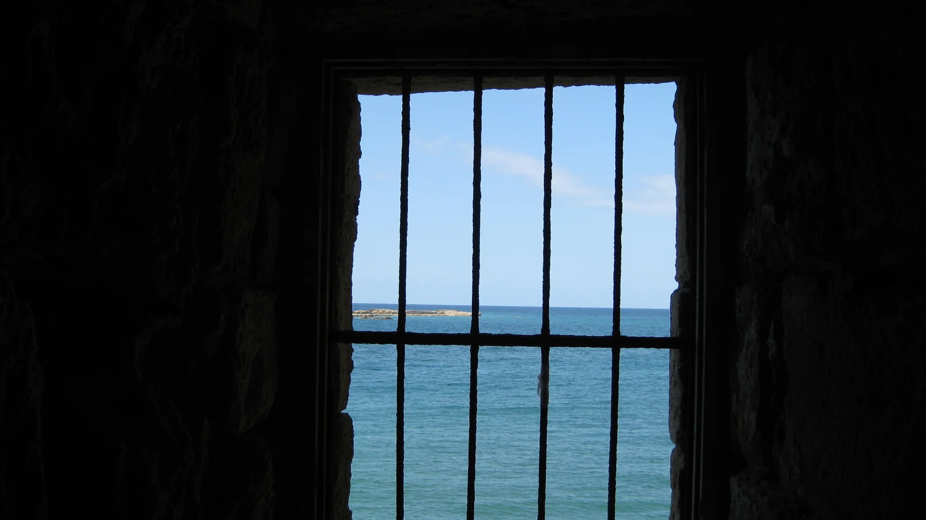 A barred rectangular window frames blue sea and sky from within a dim stone chamber.