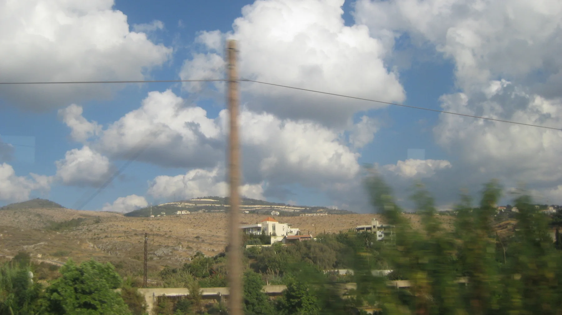 A roadside pole and wires pass before a dry hillside dotted with low buildings and greenery.