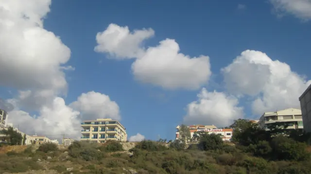 Pale apartment buildings rise above rough scrub-covered ground beneath bright clouds.