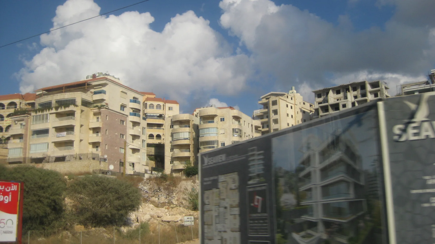 A large advertising board stands before apartment blocks on a rough sloping site.