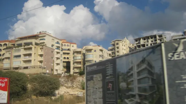 A large advertising board stands before apartment blocks on a rough sloping site.