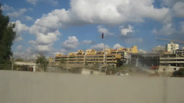 A road barrier crosses the foreground beneath partly finished buildings and a broad clouded sky.