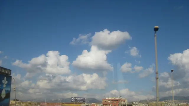 A tall street lamp rises into a blue sky filled with large white clouds above distant rooftops.